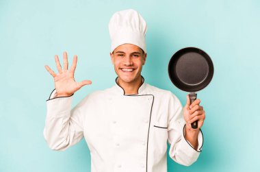 Kafkasyalı genç aşçı elinde uçan tavayla mavi arka planda izole edilmiş gülümseyen beş numarayı parmaklarıyla gösteriyor..