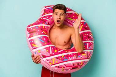 Mavi arka planda izole edilmiş şişme bir çörek tutan genç beyaz adam şaşırdı ve şok oldu..