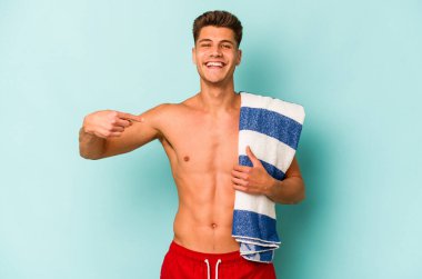Elinde plaj havlusu tutan genç beyaz adam mavi arka planda tek başına gömlek fotokopi alanını işaret ediyor, gururlu ve kendinden emin.