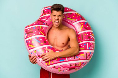 Mavi arka planda izole edilmiş şişme bir çörek tutan genç beyaz adam çok sinirli ve agresif bağırıyor..