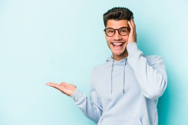 Mavi arka planda izole edilmiş genç beyaz bir adam avucunun içinde fotokopi alanı tutuyor, el üstünde tutuyor. Şaşırmış ve mutlu..