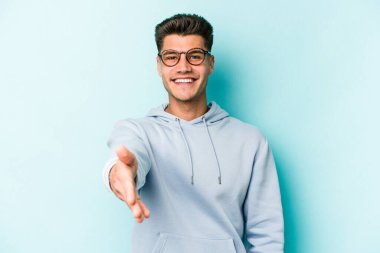 Mavi arka planda izole edilmiş genç beyaz adam selamlama jesti olarak kameraya elini uzatıyor..