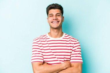 Mavi arka planda kendini güvende hisseden, kararlılıkla kollarını kavuşturan genç beyaz bir adam..