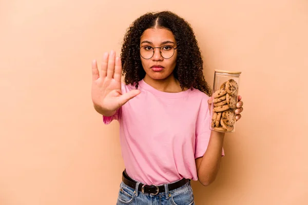 Young African american woman holding a cookies jar isolated on beige background standing with outstretched hand showing stop sign, preventing you.