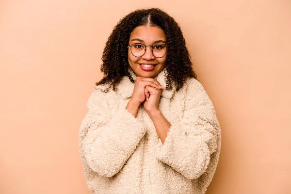 Young African American woman isolated on beige background keeps hands under chin, is looking happily aside.