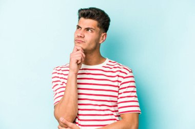 Kafkasyalı genç adam mavi arka planda izole edilmiş düşünüyor ve bakıyor, yansıtıyor, düşünüyor, bir fantezi kuruyor..