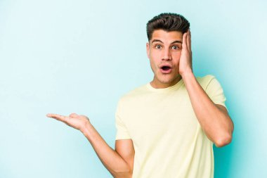 Mavi arka planda izole edilmiş genç beyaz adam, avuç içi fotokopi alanını elinde tutmaktan etkilenmiş..