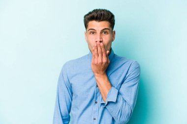 Mavi arka planda izole edilmiş genç beyaz adam şok olmuş, ağzı ellerle kaplanmış, yeni bir şey keşfetmeye hevesli..