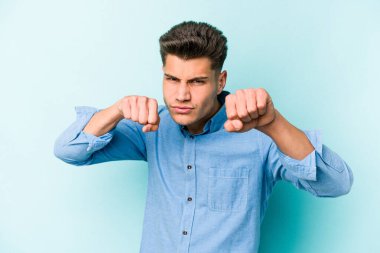 Kafkasyalı genç bir adam mavi arka planda izole edilmiş. Yumruk atıyor, sinirleniyor, kavga ediyor, kavga ediyor..