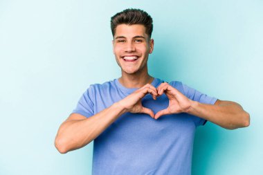 Mavi arka planda izole edilmiş genç beyaz adam gülümsüyor ve elleriyle kalp şeklini gösteriyor..