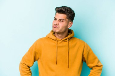 Mavi arka planda izole edilmiş genç beyaz adam omuzlarını silkeliyor ve gözleri açık..