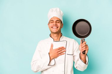 Kafkasyalı genç aşçı elinde uçan bir tavayla mavi arka planda izole bir şekilde gülüyor ve elini göğsünde tutuyor..