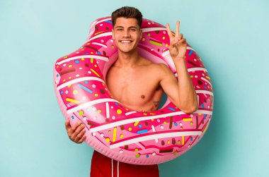 Mavi arka planda izole edilmiş şişme bir çörek tutan genç beyaz adam iki numarayı parmaklarıyla gösteriyor..