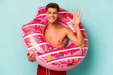 Genç beyaz adam mavi arka planda izole edilmiş şişme bir çörek tutuyor. Beş numarayı parmaklarıyla gülümsüyor..