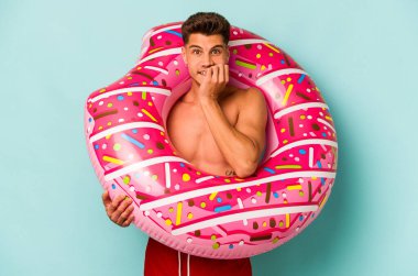 Mavi arka planda izole edilmiş, tırnak yiyen, gergin ve endişeli, şişme donut tutan genç beyaz bir adam..