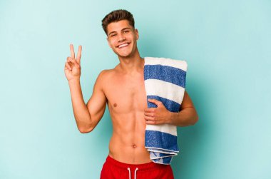 Elinde plaj havlusu tutan genç beyaz adam mavi arka planda izole edilmiş neşeli ve tasasız parmakları olan bir barış sembolü gösteriyor..