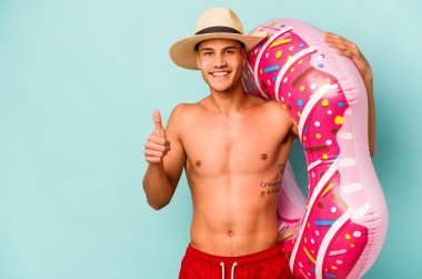 Genç beyaz adam mavi arka planda izole edilmiş şişme bir çörek tutuyor gülümsüyor ve başparmağını kaldırıyor.