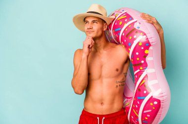 Mavi arka planda izole edilmiş şişme donut tutan beyaz bir genç şüpheli ve şüpheci bir ifadeyle yan yana bakıyor..