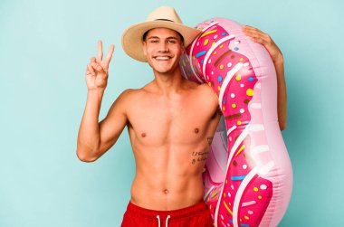 Mavi arka planda izole edilmiş şişme bir çörek tutan genç beyaz adam iki numarayı parmaklarıyla gösteriyor..