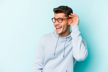 Mavi arka planda izole edilmiş genç beyaz adam bir dedikoduyu dinlemeye çalışıyor..