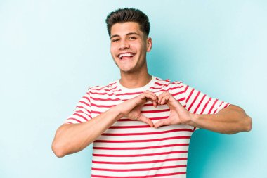 Mavi arka planda izole edilmiş genç beyaz adam gülümsüyor ve elleriyle kalp şeklini gösteriyor..