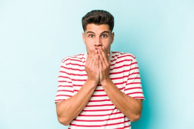 Mavi arka planda izole edilmiş genç beyaz adam şok olmuş, ağzı ellerle kaplanmış, yeni bir şey keşfetmeye hevesli..