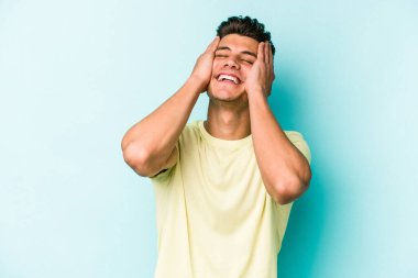 Mavi arka planda izole edilmiş genç beyaz adam ellerini başının üstünde tutarak neşe içinde gülüyor. Mutluluk kavramı.