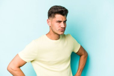 Kafkasyalı genç adam mavi arka planda izole edilmiş hoşnutsuzca kaşlarını çatmış, kollarını kavuşturmuş..