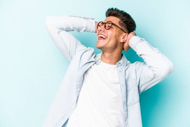 Kafkasyalı genç adam mavi arka planda izole edilmiş. Kendine güveniyor, elleri başının arkasında..