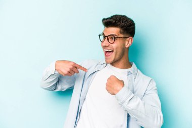 Mavi arka planda izole edilmiş genç beyaz adam parmağını göstererek şaşırdı, geniş bir gülümsemeyle..