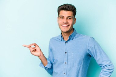 Mavi arka planda izole edilmiş genç beyaz adam işaret parmağıyla işaret ederek gülümsüyor..