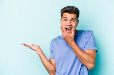 Mavi arka planda izole edilmiş genç beyaz bir adam avucunun içinde fotokopi alanı tutuyor, el üstünde tutuyor. Şaşırmış ve mutlu..