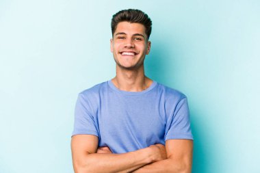 Mavi arka planda kendini güvende hisseden, kararlılıkla kollarını kavuşturan genç beyaz bir adam..