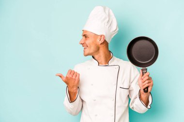 Kafkasyalı genç aşçı elinde uçan tavayla mavi arka planda izole edilmiş, başparmak uzakta, gülerek ve kaygısızca.