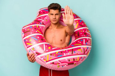 Mavi arka planda izole edilmiş şişirilebilir donut tutan genç beyaz bir adam elini uzatmış, dur işareti göstererek seni engelliyor..