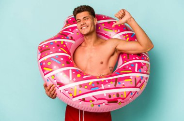 Young caucasian man holding an inflatable donut isolated on blue background feels proud and self confident, example to follow.
