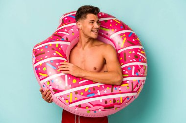 Mavi arka planda izole edilmiş şişme bir çörek tutan genç beyaz adam gülümsüyor, neşeli ve keyifli görünüyor..