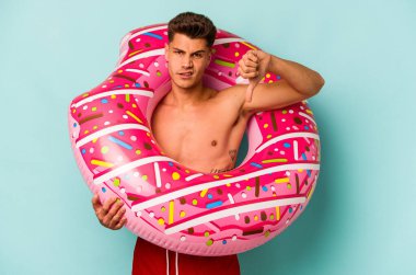 Mavi arka planda izole edilmiş şişme donut tutan genç beyaz bir adam, hoş olmayan bir jest gösteriyor, kabul etmiyor. Anlaşmazlık konsepti.