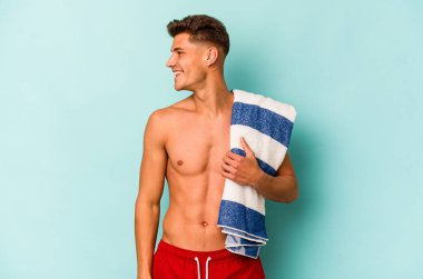 Mavi arka planda izole edilmiş plaj havlusu tutan beyaz bir adam gülümsüyor ve keyifli görünüyor..