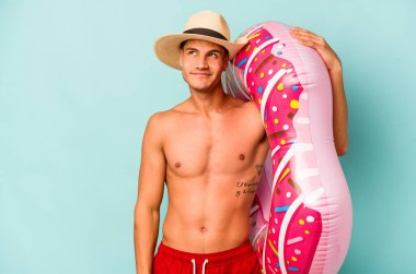 Kafkasyalı genç adam mavi arka planda izole edilmiş şişirilebilir bir çörek tutuyor. Amaçlarına ve amaçlarına ulaşma hayalleri kuruyor.