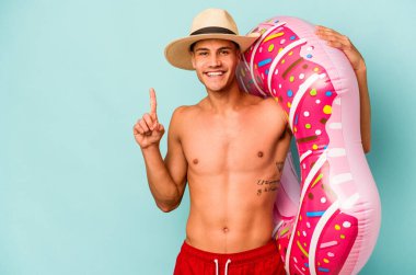 Mavi arka planda izole edilmiş şişirilebilir donut tutan genç beyaz bir adam bir numarayı parmağıyla gösteriyor..