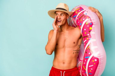 Mavi arka planda izole edilmiş şişme donut tutan genç beyaz adam gizli bir sıcak fren haberi veriyor ve yan bakıyor.