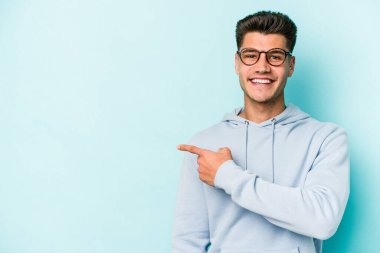 Mavi arka planda izole edilmiş genç beyaz adam gülümsüyor ve boş alanda bir şeyler gösteriyor..