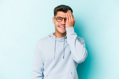 Mavi arka planda izole edilmiş genç beyaz adam yüzünün yarısını palmiye ile kaplayarak eğleniyor..