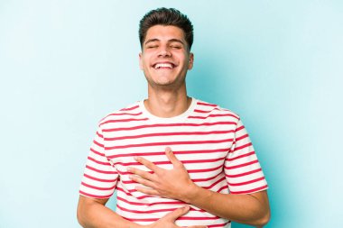 Mavi arka planda izole edilmiş genç beyaz adam mutlu bir şekilde gülüyor ve ellerini karnının üstünde tutarak eğleniyor..