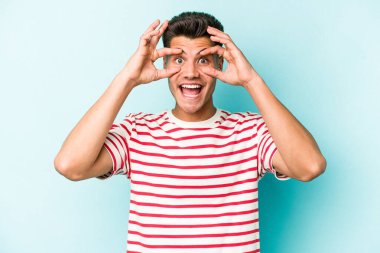 Mavi arka planda izole edilmiş genç beyaz adam bir başarı fırsatı bulmak için gözlerini açık tutuyor..