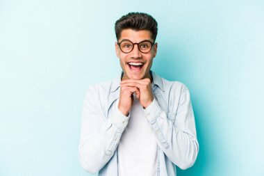 Kafkasyalı genç adam mavi arka planda izole edilmiş şans için dua ediyor, şaşırmış ve ağzını açıp önüne bakıyor..