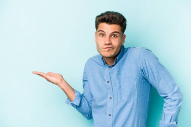 Mavi arka planda izole edilmiş genç beyaz bir adam bir palmiye üzerinde fotokopi alanı ve belinde bir el daha gösteriyor..