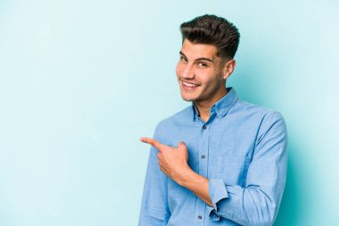 Mavi arka planda izole edilmiş genç beyaz adam gülümsüyor ve boş alanda bir şeyler gösteriyor..