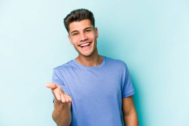Mavi arka planda izole edilmiş genç beyaz adam selamlama jesti olarak kameraya elini uzatıyor..